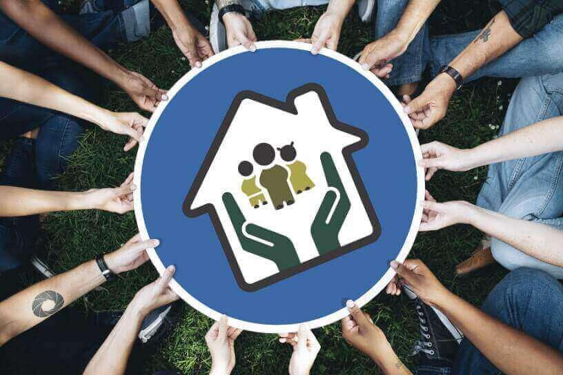 Eine gemischte Gruppe sitzt im Kreis auf einer Wiese und hält ein großes rundes Schild hoch. Darauf ist ein Haus mit einer Familie darin abgebildet. Unter dem Schild sind Hände zu sehen, die Schutz und Unterstützung der Gemeinschaft symbolisieren.